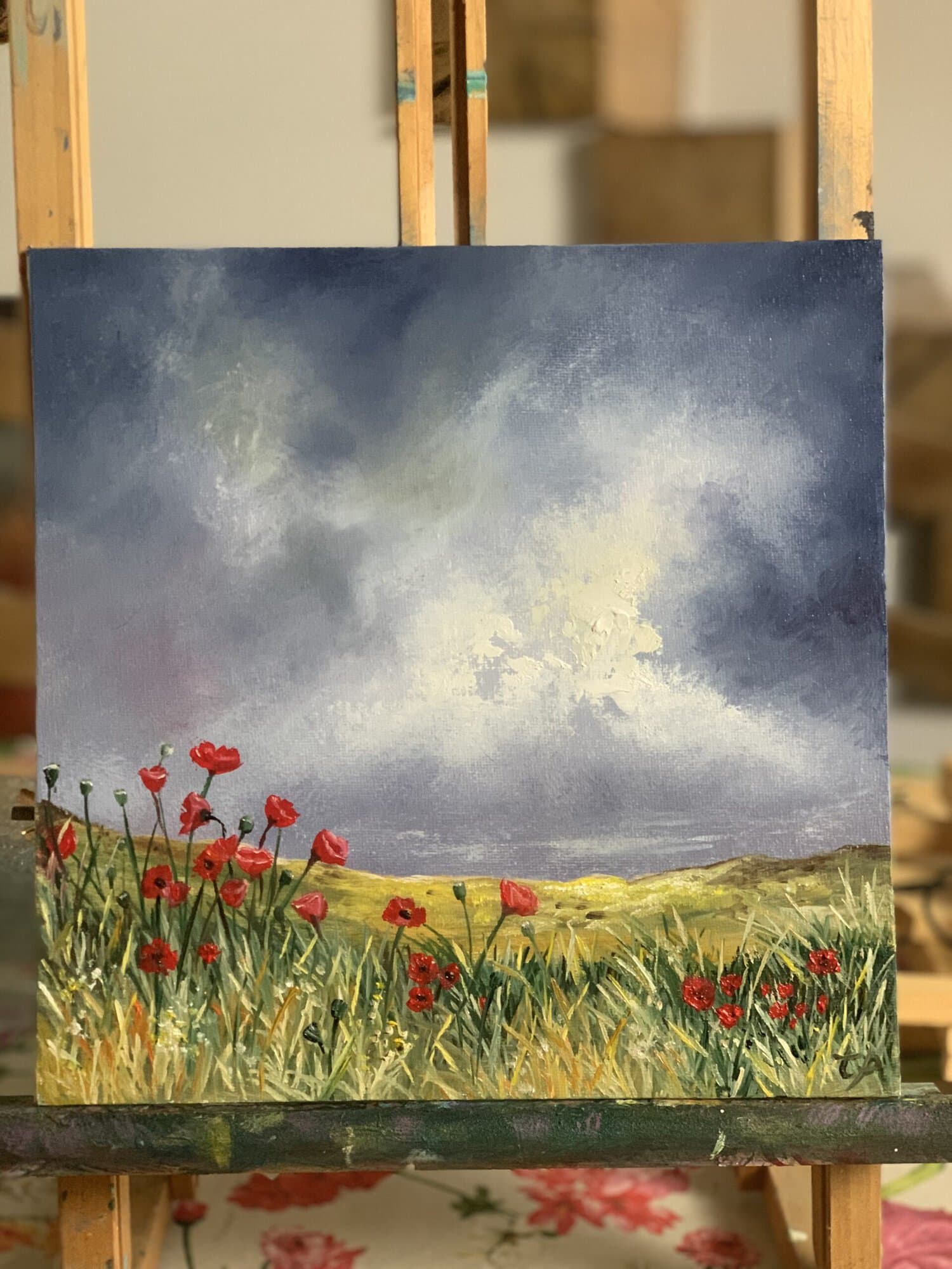 Original 25 × 25 cm oil landscape painting featuring vibrant red poppies beneath dramatic storm clouds with sunlight breaking across rolling countryside in rural France, painted in an expressive contemporary style.